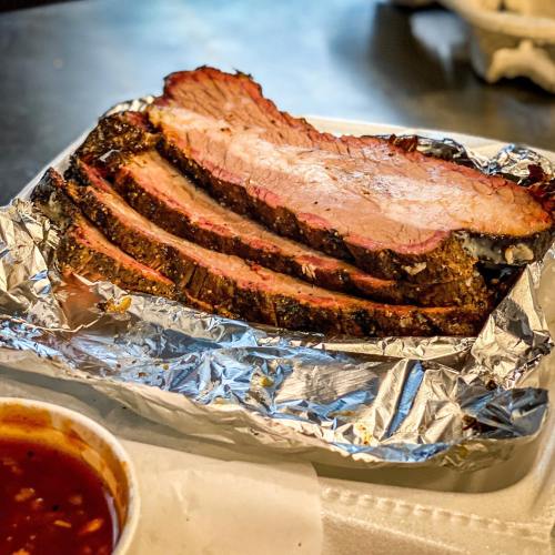 Sliced smoked brisket on a foil tray with a side of barbecue sauce.