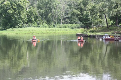 Two kayakers paddle on a calm lake surrounded by lush greenery and trees, with a dock visible in the background.