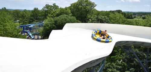A person rides a large inflatable tube down a water slide surrounded by trees and a clear blue sky.