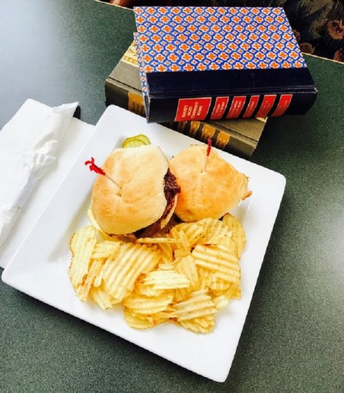 A plate with two sliders topped with pickles, served with a side of wavy potato chips and stacked books in the background.