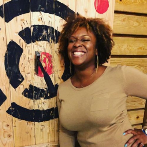 A smiling woman with curly hair stands next to a wooden wall with a circular design. She wears a beige sweater.