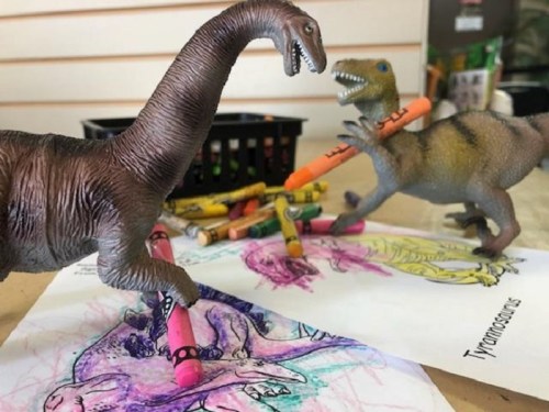 Two toy dinosaurs, a long-necked dinosaur and a T. rex, are playfully holding crayons over colorful dinosaur drawings.