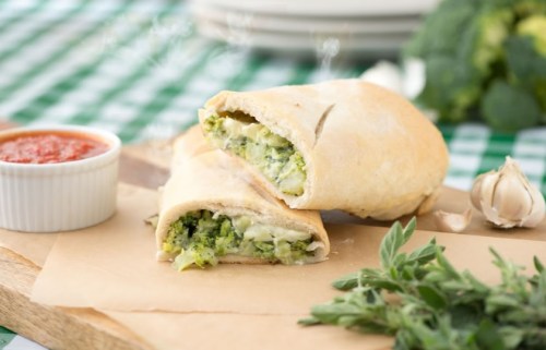 A cut calzone filled with broccoli and cheese, served with marinara sauce and fresh herbs on a wooden board.