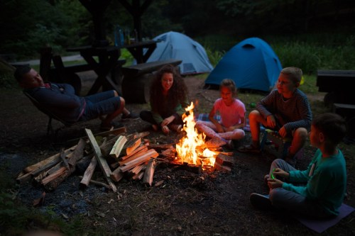 A group of people sitting around a campfire at night, with tents and trees in the background.