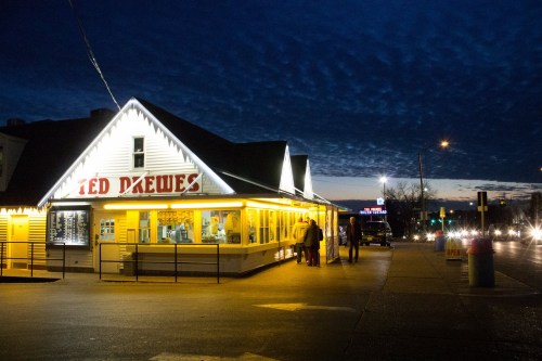 A brightly lit ice cream shop at dusk, with people outside and a cloudy sky in the background.