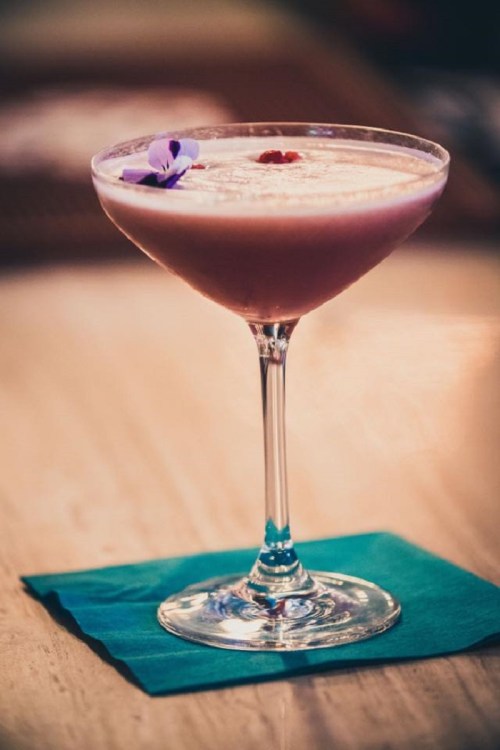 A pink cocktail garnished with a flower, served in a elegant glass on a teal napkin.