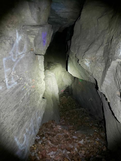 A narrow, dimly lit passage between large rock formations, with faint graffiti and fallen leaves on the ground.