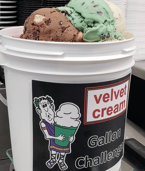 A bucket filled with various ice cream flavors, featuring a cartoon character holding ice cream and a "Gallon Challenge" sign.