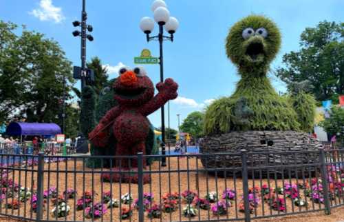 Colorful topiary figures of Elmo and a green monster in a park, surrounded by flowers and trees.