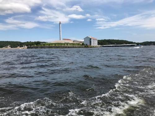 A view of a lake with waves, featuring a building and a tall smokestack in the background under a partly cloudy sky.