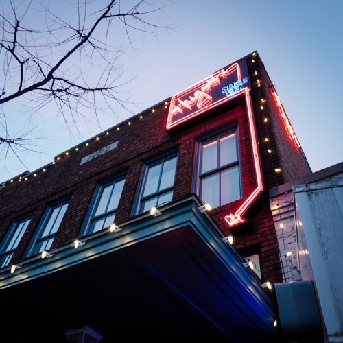 Neon sign on a brick building, with decorative lights and a clear blue sky in the background.