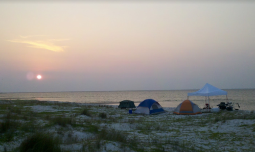 Tents and a canopy set up on a beach at sunset, with calm waters and a soft sky in the background.