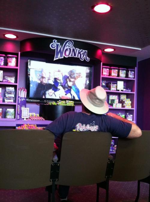 A person in a hat sits in front of a TV in a colorful shop filled with candy, labeled "Wonka."