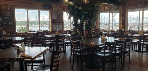 Interior of a restaurant with wooden tables and chairs, large windows overlooking a marina with boats.