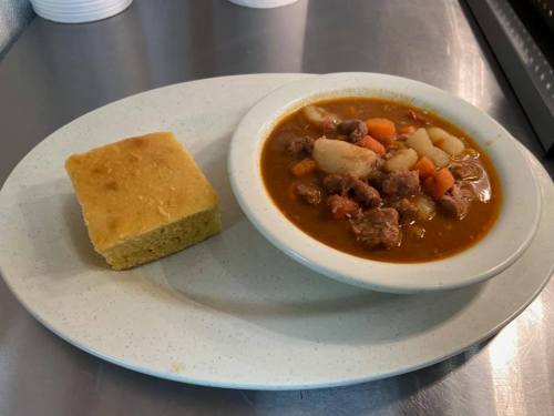 A bowl of hearty meat and vegetable stew served with a slice of cornbread on a white plate.