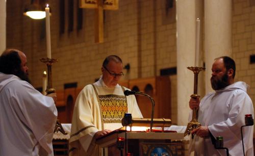 A priest stands at a lectern with two altar servers holding candles in a dimly lit church.