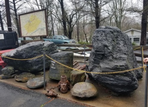 Two large rocks displayed outdoors, surrounded by a chain, with a map and vehicles in the background on a rainy day.
