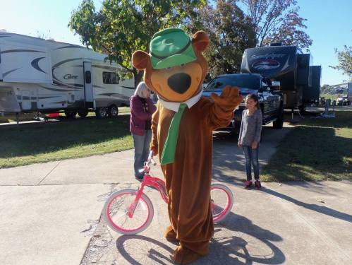A bear mascot waves beside a pink bicycle, with people and RVs in the background on a sunny day.
