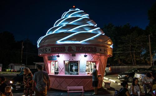 A brightly lit ice cream stand shaped like a giant ice cream cone, with people gathered outside at night.