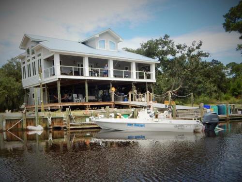 A white house on stilts overlooking a calm waterway, with a boat docked nearby and trees in the background.