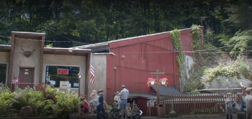 A rustic building with a red exterior, surrounded by greenery, and people gathered outside.