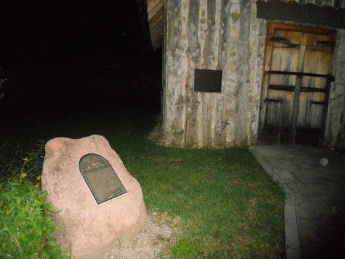 A stone monument with a plaque stands near a rustic wooden building at night, surrounded by grass and plants.