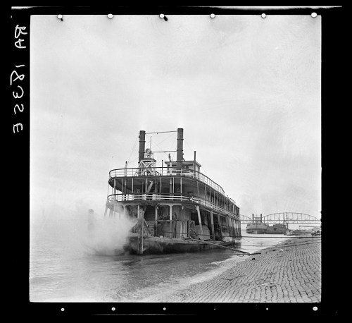 A vintage steamboat docked by the shore, with smoke billowing from its stacks and a bridge in the background.