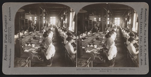 Workers in a jewelry manufacturing workshop, seated at long tables, focused on their tasks in a historic setting.