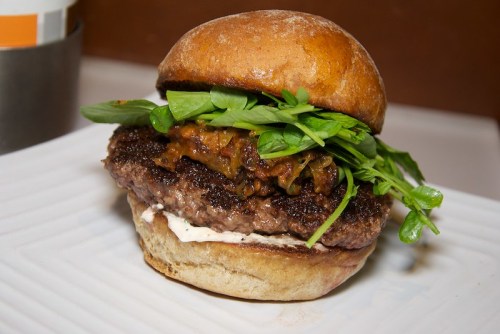A juicy burger with a toasted bun, topped with greens and a flavorful sauce, served on a white plate.