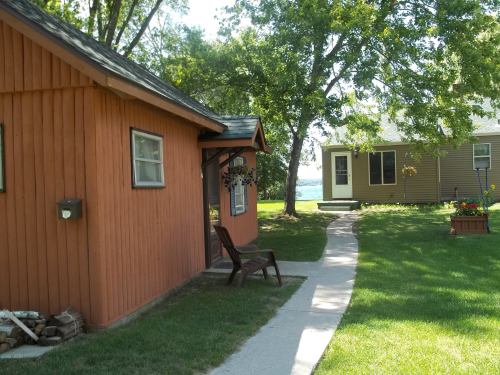 A pathway leads between two houses surrounded by green grass and trees, with a chair on the porch of the orange house.