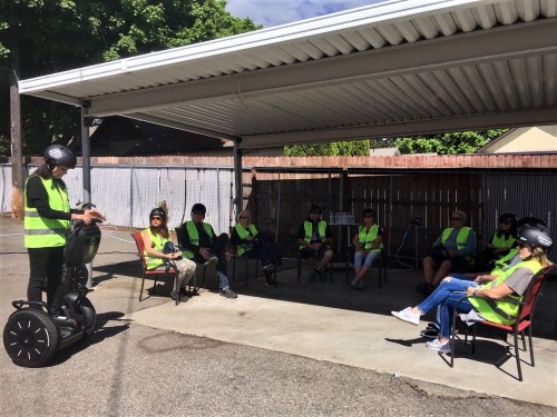 A group of people in safety vests and helmets sits in chairs while one person stands on a Segway, speaking to them.