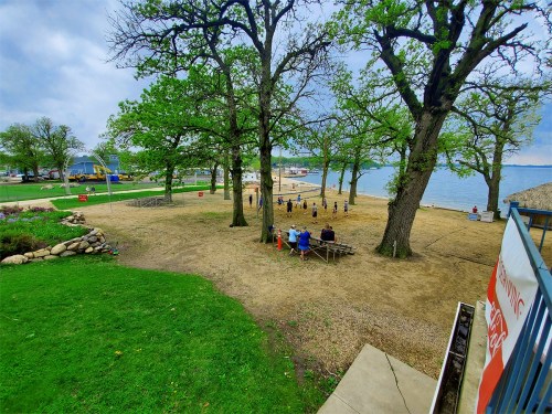 A beach area with trees, people playing volleyball, and a view of the water under a cloudy sky.