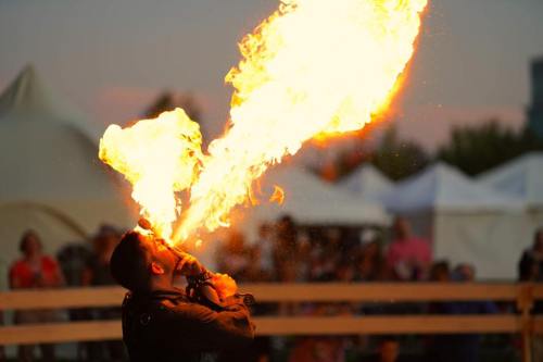 A performer breathes fire, creating a dramatic flame against a twilight sky, with an audience in the background.