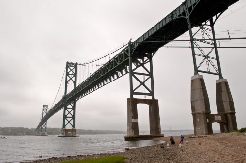 A large bridge spans over a river, with two people walking along the shore under a cloudy sky.