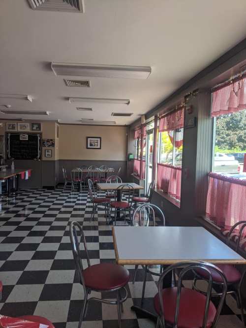 A cozy diner interior with checkered floors, red and white decor, and empty tables by large windows.