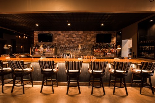 A stylish bar with a wooden counter, high stools, and a wall of shelves filled with various bottles.