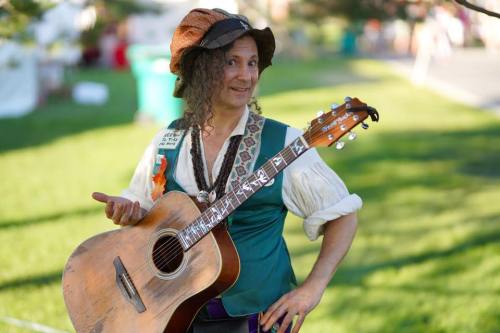 A musician in a colorful outfit holds an acoustic guitar, smiling and posing outdoors in a park setting.