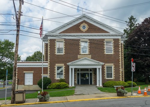 A brick building with columns, an American flag, and a sign, surrounded by greenery and power lines.