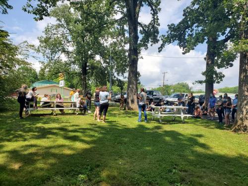 A group of people gathered outdoors around picnic tables under trees on a sunny day.