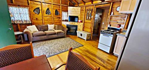 Cozy wooden cabin interior with a living area, kitchen, and dining table, featuring a sofa and fireplace.