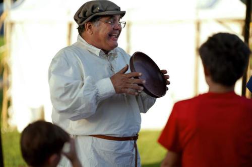 A smiling man in historical clothing holds a plate while engaging with children at an outdoor event.