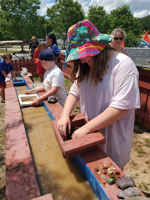 Children and adults participate in a hands-on activity, sifting through water and rocks at a mining station outdoors.