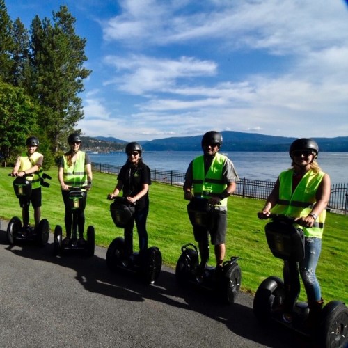 A group of five people in safety vests riding segways along a scenic waterfront path with trees and mountains in the background.