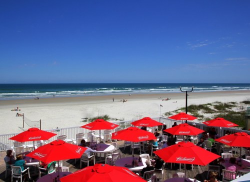 A beachside view with red umbrellas over tables, overlooking a sandy shore and calm ocean under a clear blue sky.