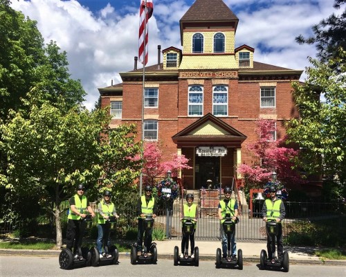 A group of six people in vests stand on Segways in front of Roosevelt School, surrounded by trees and flowers.
