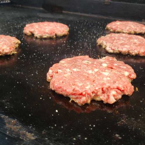 Five raw hamburger patties cooking on a griddle, with seasoning visible on the surface.