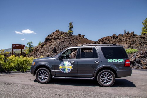 A gray SUV parked near a sign for North Crater 2, with volcanic rock formations in the background and blue sky above.