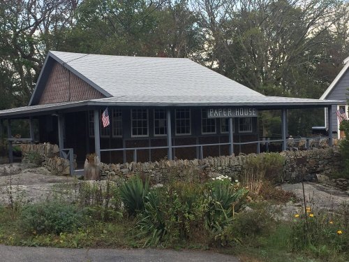 A rustic house with a porch, surrounded by greenery and stone walls, featuring an American flag.