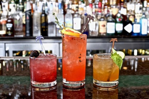 Three colorful cocktails on a bar counter, garnished with fruit and herbs, against a backdrop of liquor bottles.