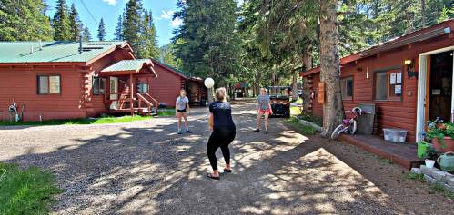 A group of people playing volleyball in a forested area with cabins and trees in the background.